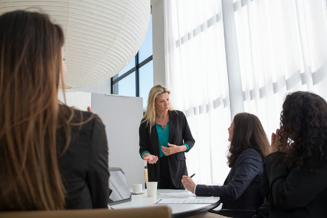 Profissional conduzindo uma reunião de equipe em pé, com o time sentado à mesa em uma sala de reuniões.